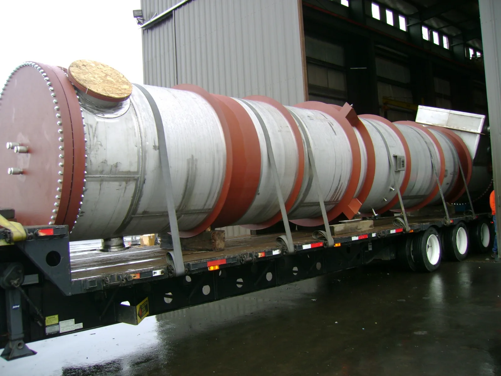Large fabricated stainless steel vessel with bolted head and primed support rings, loaded on a flatbed trailer for shipment